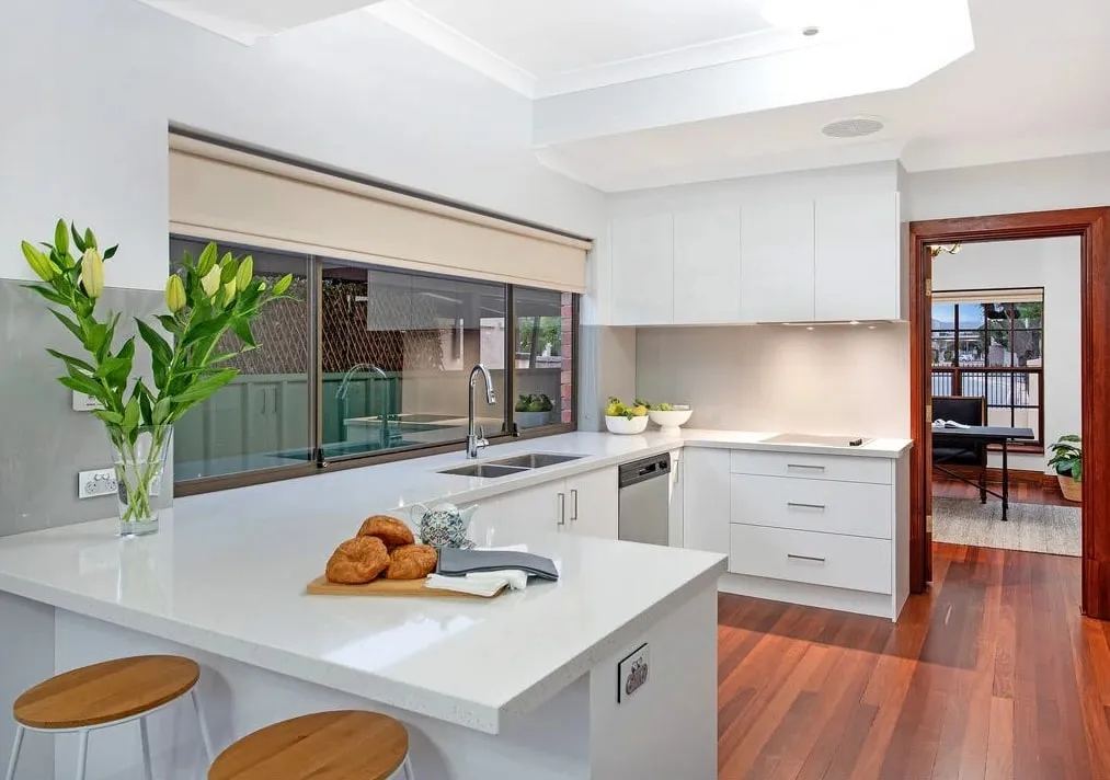 A white U shaped kitchen with a stone benchtop, a window over the sink and timber floors
