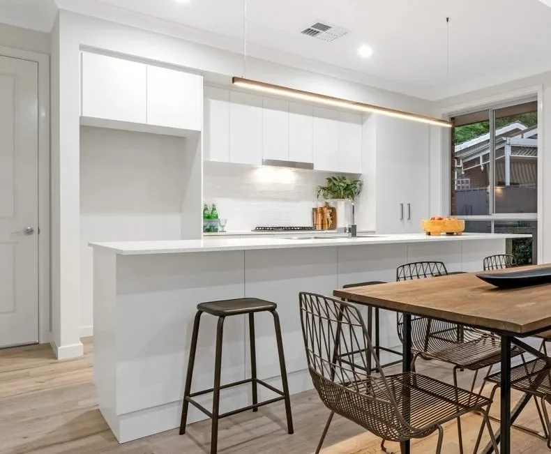 A white handleless kitchen with a long island, a linear pendant light and a timber dining table